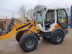 Gelber und schwarzer Radlader steht im Dreck, umgeben von Erde und Baustellenmaterial.