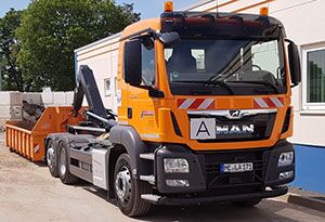 Ein Containerfahrzeug in orange steht mit heruntergelassenem Container vor einem Gebäude.